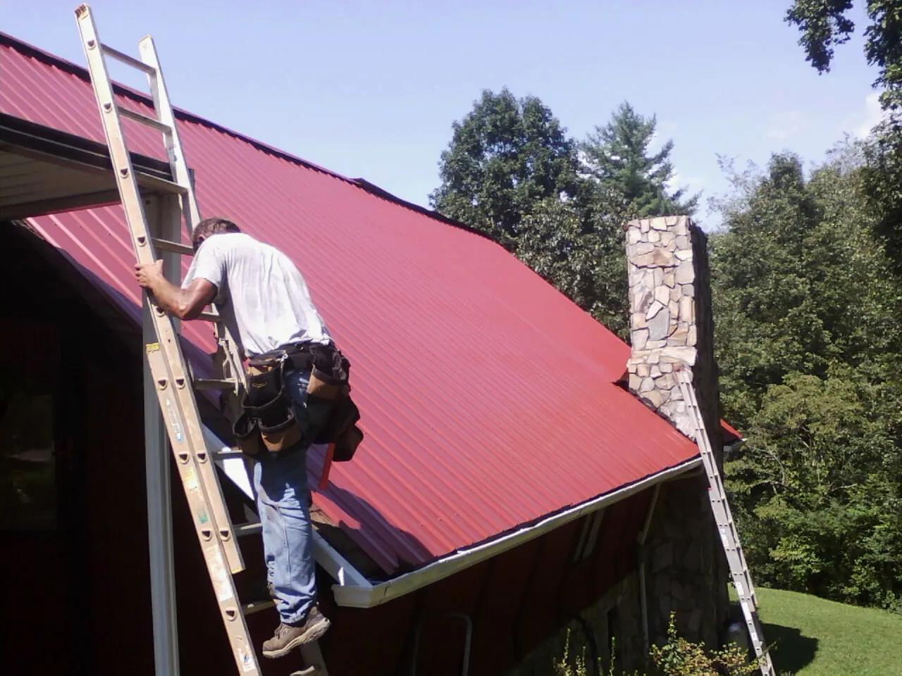 Roofer on a ladder beside a red metal roof and stone chimney — Soffit & Fascia Repair by Roofing Brothers in Metter